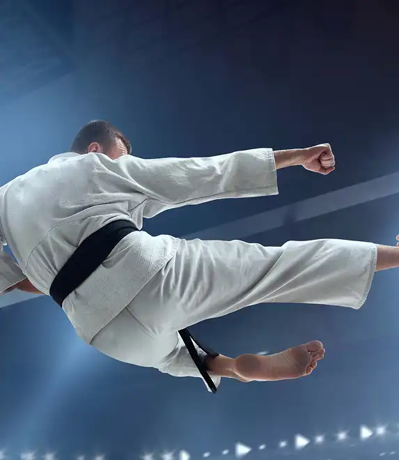 Un artiste martial vêtu d'un gi blanc exécute un coup de pied dans les airs dans une arène éclairée par des projecteurs. Le décor traduit le mouvement dynamique et la concentration.